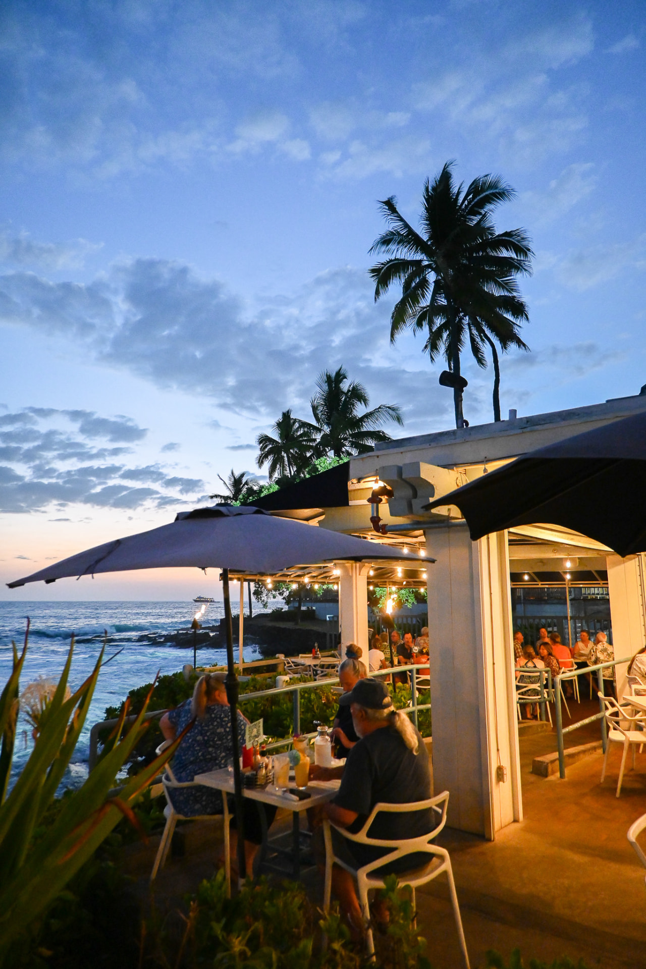 Guests dining under umbrellas on the Kai lanai at sunset above the surf